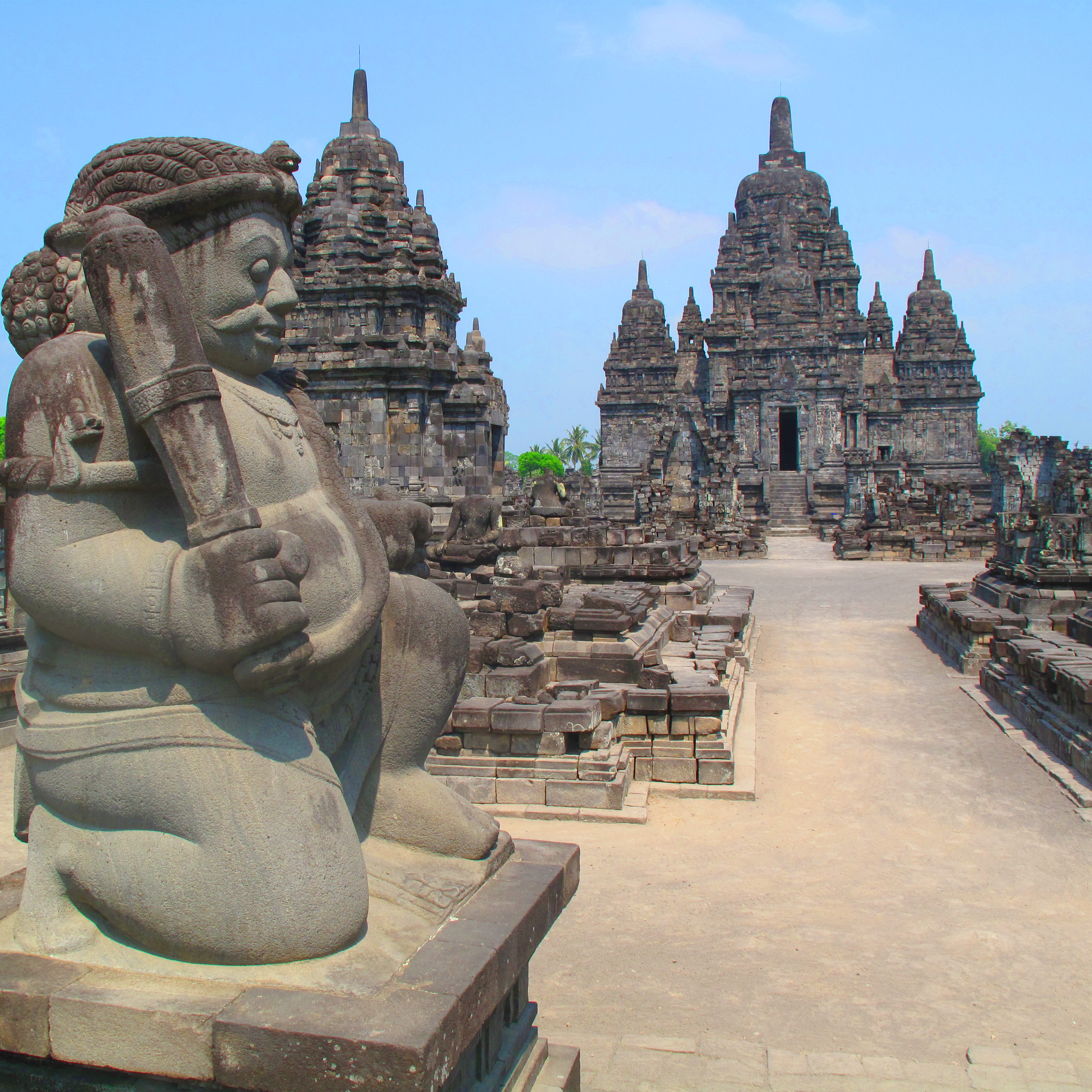 Carved gray statue of a stout man with a club. In the background are templa structures of the same stone.