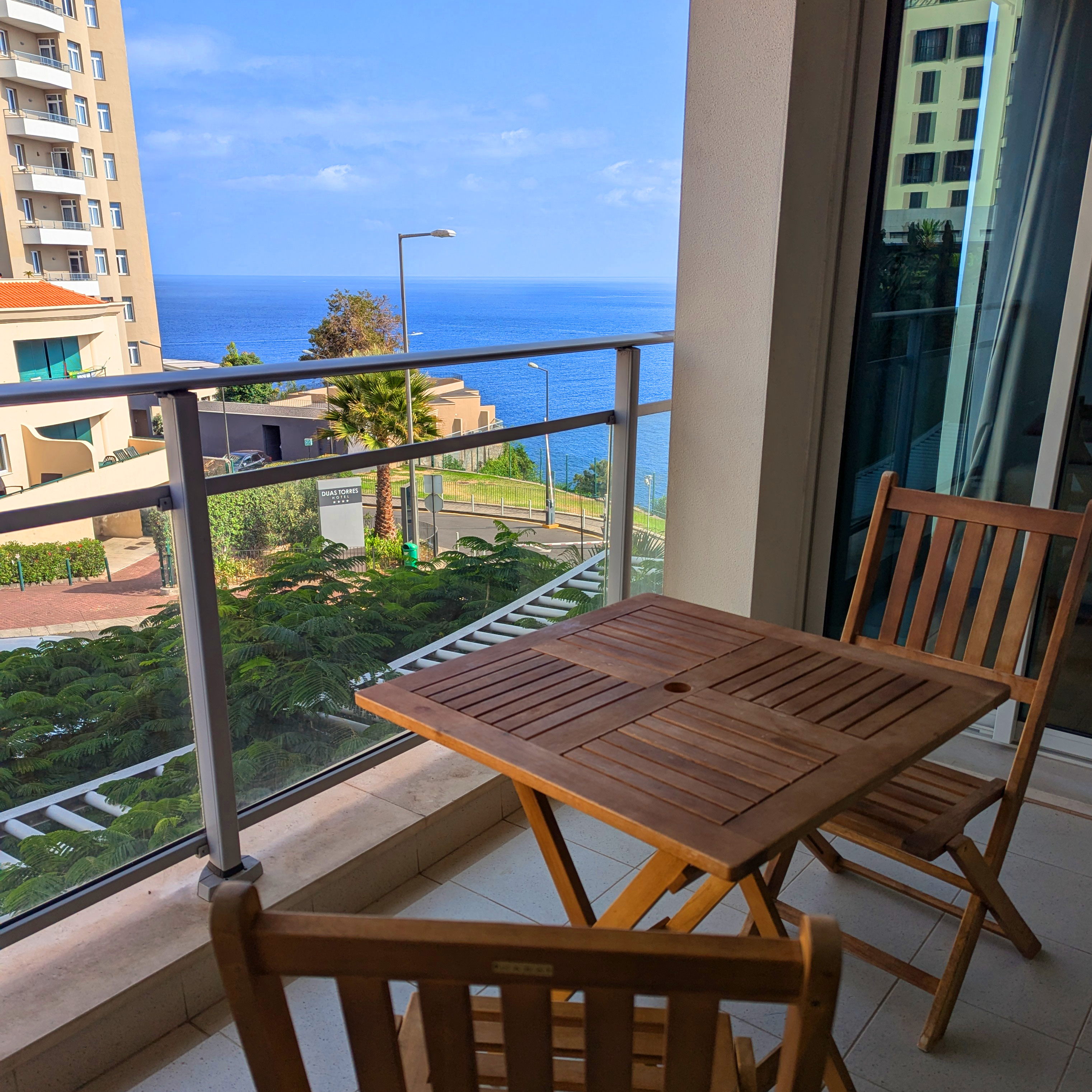 A wooden slat table and chair sit in the foreground, with a view of the ocean beyond the railing.