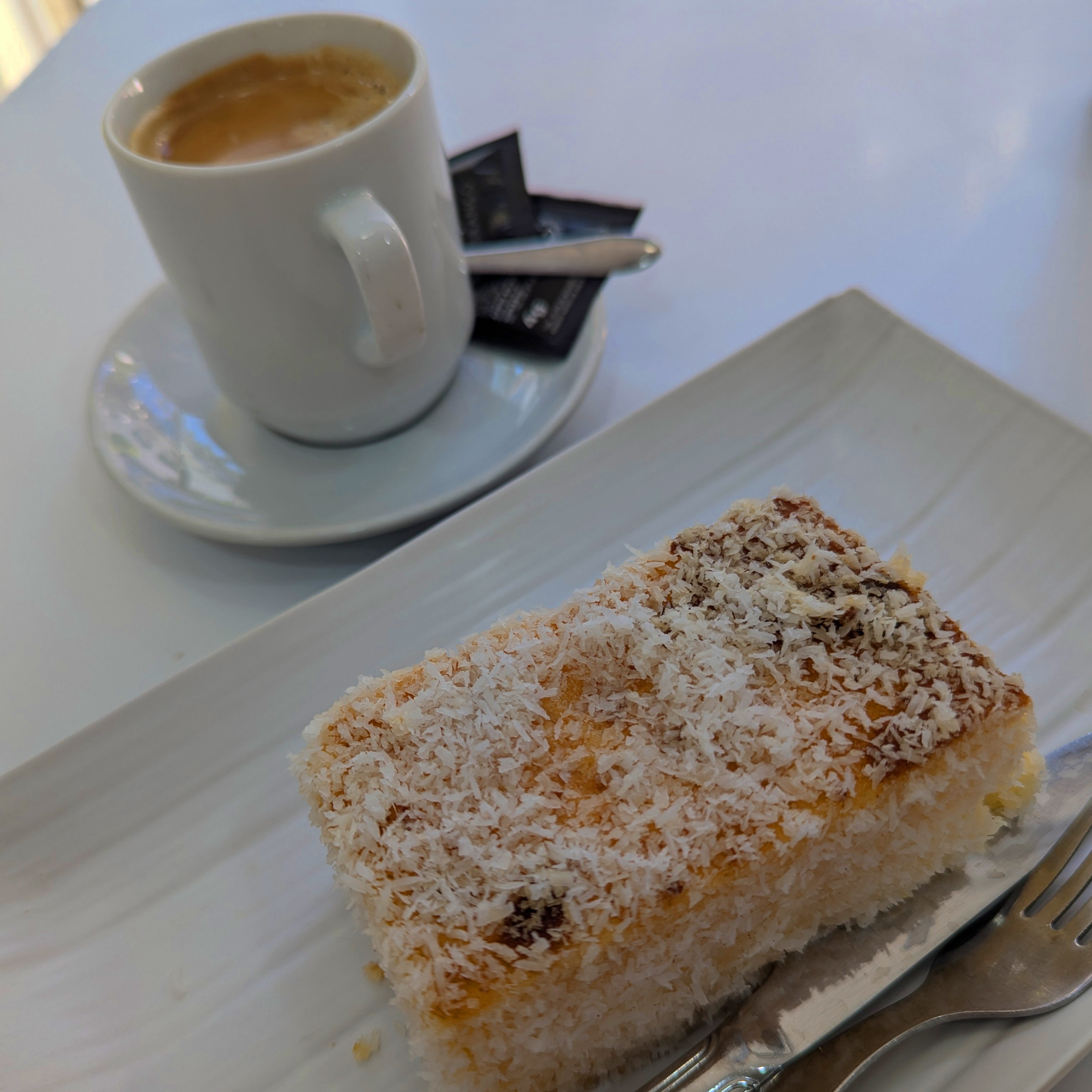 A rectangular slice of cake covered in coconut with a double expresso in the background.