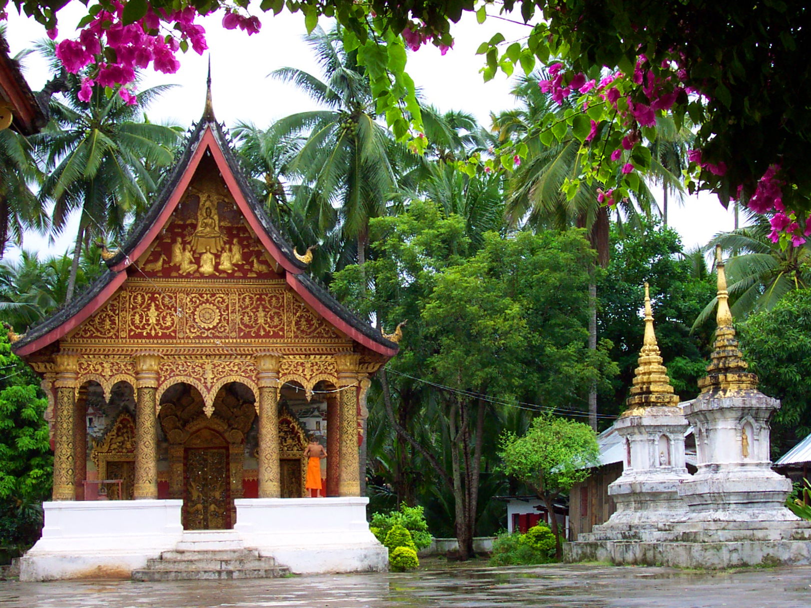 Peering through some flowering bushes at a small temple building decorated with red and gold. A monk is adjusting his robe on the porch of the building. To the left are two funerary monuments.