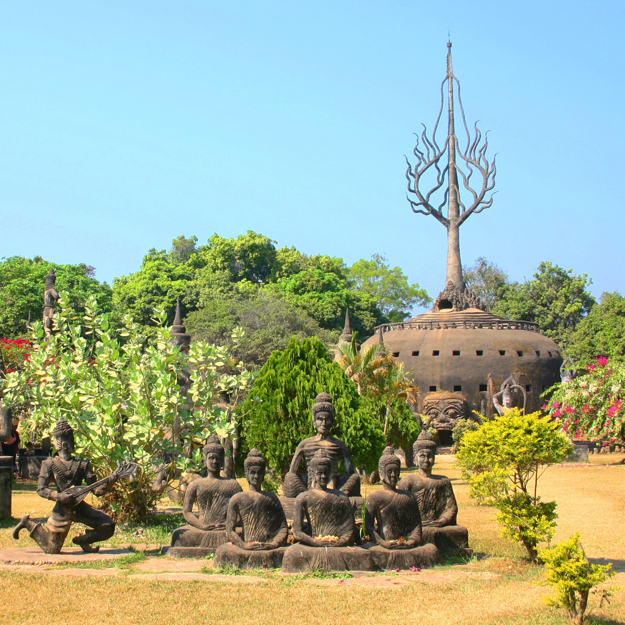 View of Buddha statues. In the background is a large pumpkin-shaped building with a sort of tree sprouting from the top and a doorway framed by a demon head.